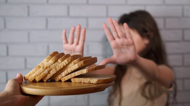 Young girl refusing bread expressing gluten intolerance. Unhappy teenage girl showing a stop gesture with her hands to a plate of sliced bread, symbolizing a gluten-free diet, celiac disease awareness
