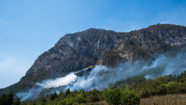 Firefighting aircraft engaged in putting out a large forest fire on a rocky slope, characterised by thick smoke and high-altitude water drops