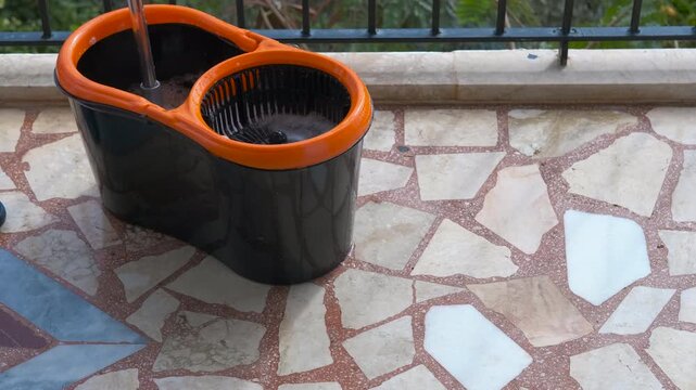 Housekeeper wringing mop in bucket on balcony. Professional housekeeper cleaning balcony floor, using orange and black bucket with mop and wringer, ensuring thorough removal of dirt