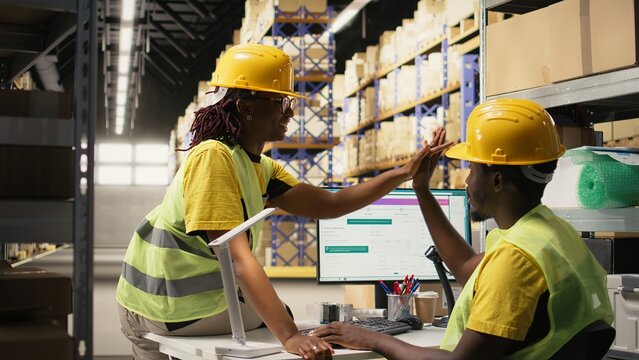 Black staff members sharing happy high five gesture in busy storehouse setting, proud and pleased with parcel shipment teamwork success.