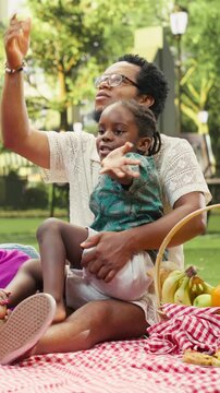Vertical video: Father and mother point toward the sky while a curious boy looks up, smiling together during an outdoor picnic in an urban park and sharing family bonding and leisure on a sunny day.