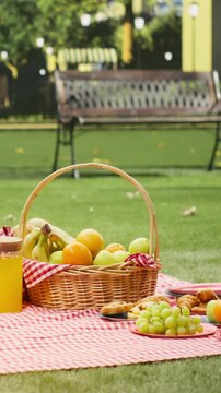 Portrait format: Picnic basket is filled with fresh fruits and orange juice on a blanket in a public park, with apples, oranges, bananas and grapes arranged as picnic food.