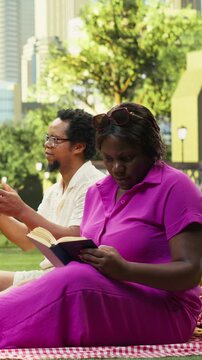 Portrait format: Mother reads a book on a picnic blanket while the boy and dad play with a soccer ball nearby, enjoying weekend fun and relaxation in a city park.