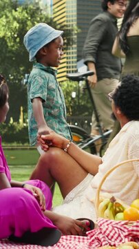Vertical aspect: Expressive boy gestures while talking to parents at picnic as the family shares playful conversation beside fruit basket and food in public park, during leisure and family bonding.