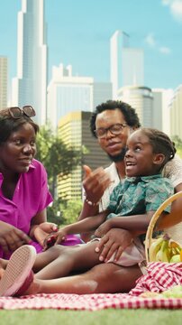 Vertical frame: Happy parents share a playful conversation with their boy while relaxing beside picnic food on a checkered blanket in an urban park.