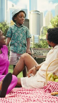 Vertical frame: Joyful boy stands between seated parents and speaks during picnic, while the family enjoys weekend fun together beside food basket in sunny urban park.