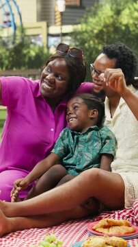 Vertical composition: Joyful parents and boy cuddle for photos while relaxing on a picnic blanket with food and a city skyline behind them during family bonding and weekend fun.
