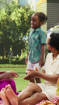 Vertical video: Young boy dances and has fun with his parents on a checkered blanket, fooling around and laughing with silly moves during a picnic and leisure beside a fruit basket in an urban park.