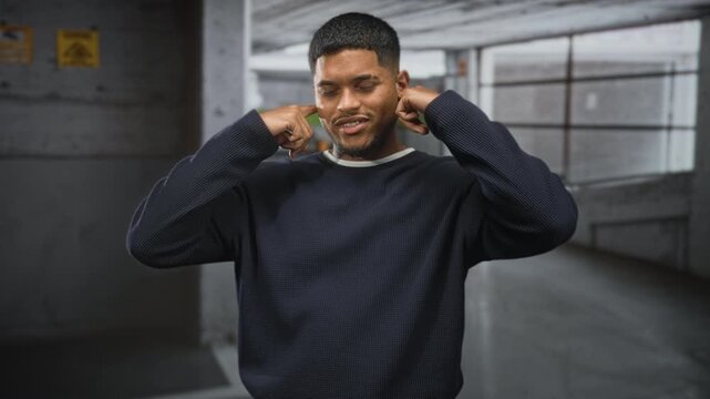 Man plugs ears with fingers and grimaces while standing in a dim concrete parking building, wearing a dark sweater and beard visible; annoyance.