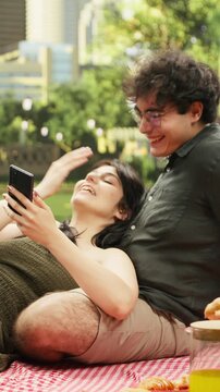 Vertical frame: Laughing young woman leans on a man and uses a smartphone while they share amusing reels on a picnic blanket in an urban park, creating a funny moment together during their date.