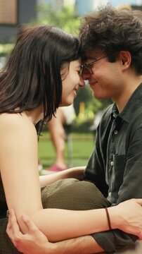 Vertical screen: Couple touch foreheads with an intimate gaze while sitting together on a picnic blanket in a city park and showing romantic affection face to face.