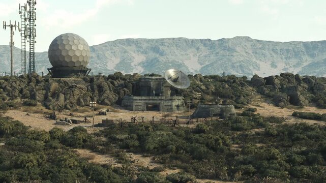 A communication facility sits on rocky terrain, featuring large satellite dishes and antennas surrounded by sparse vegetation and distant mountains under a clear sky.
