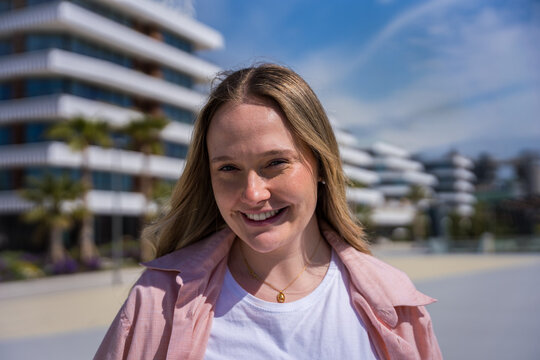 Young blonde woman smiles directly at the camera while standing outdoors in a modern urban neighborhood on a bright sunny day, conveying confidence and positivity.
