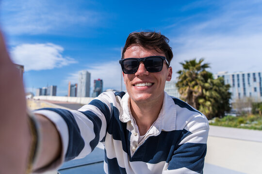 Young man in sunglasses smiles while taking a selfie outdoors in a contemporary urban area, sitting casually under a bright blue sky near modern buildings and palm trees.