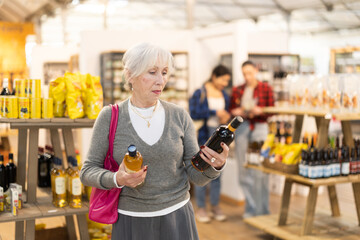 Fototapeta premium Elderly woman buyer chooses bottle of wine in grocery store