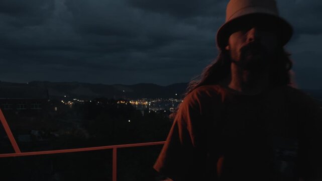 Solitary long-haired man in bucket hat stands on dark terrace leaning on railing looking at night city lights then turns and walks towards camera with face visible under dramatic cloudy dusk sky