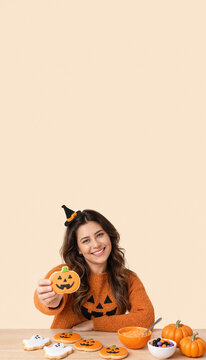 Smiling woman holding pumpkin shaped cookie at wooden table with Halloween treats and decor