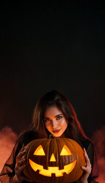 Portrait of beautiful girl holding illuminated jack-o-lantern pumpkin with smoke on dark background