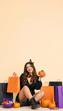 Smiling young woman in witch headband holding pumpkin with colorful shopping bags on floor