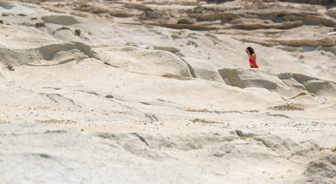A woman walks across pale rock formations near Mandrakia, Milos, Greece, surrounded by sunlit coastal terrain.