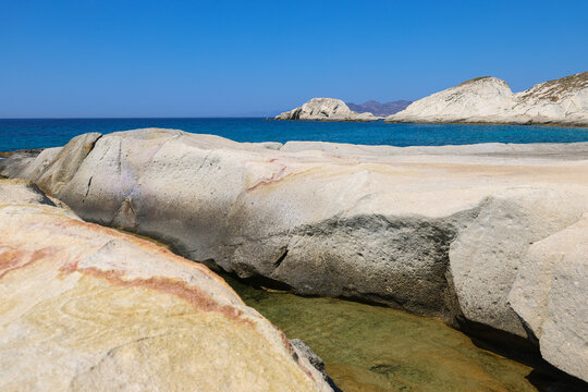 Smooth white rocks meet turquoise water along the coast near Mandrakia, Milos, Greece, under a clear summer sky.