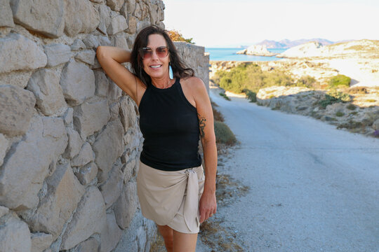 A woman leans against a stone wall overlooking the coastal landscape near Mandrakia, Milos, Greece, under warm afternoon light.