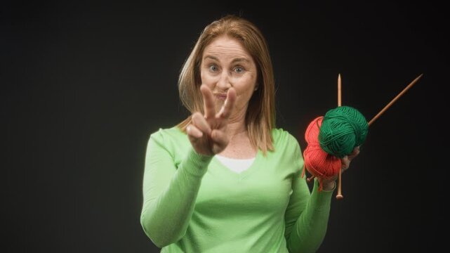 Woman wearing green sweater holding green and red yarn with wooden knitting needles, holding up two fingers toward camera in studio; playful charm.