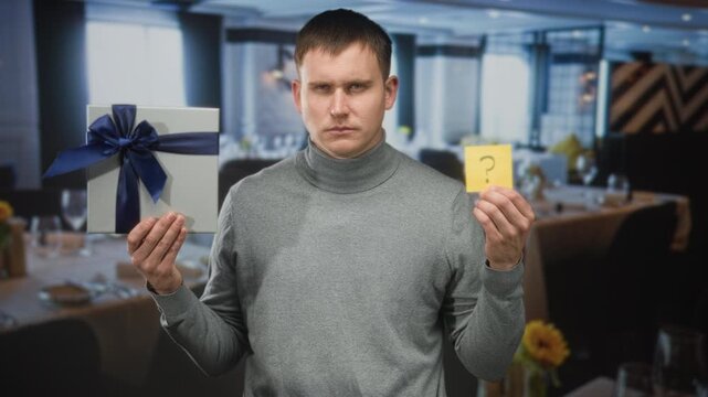 Man holds gift box and shows question mark note with bare hands in a building dining room; uncertainty decision choice.