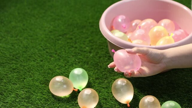 Woman taking water balloon from basin, tossing and squeezing it on green grass, closeup
