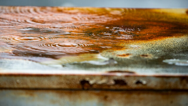 Raindrops falling on a rusty metal surface creating ripples close up.A beautiful high-contrast close-up shot capturing heavy raindrops falling onto a weathered