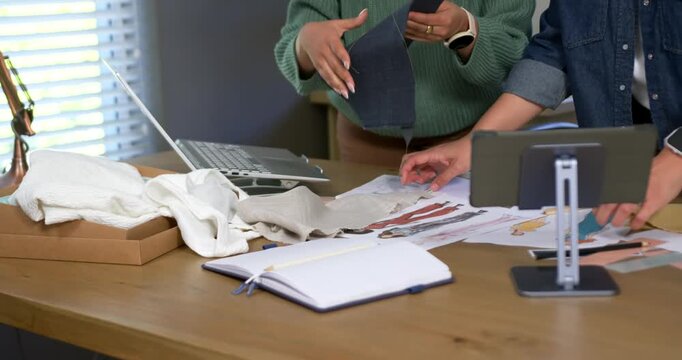 Asian, African American women choosing swatch and laying fabrics over sketches at table for design
