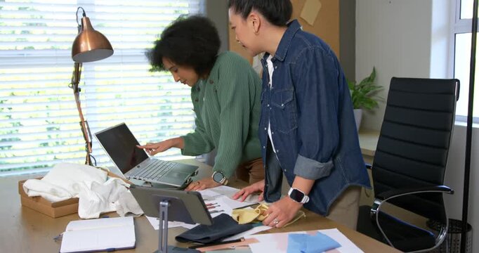 Diverse women designers in green sweater denim shirt using laptop and comparing samples, deciding
