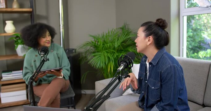 Diverse women hosts starting podcast in studio holding tablet, guest speaking, fixing mic, smiling