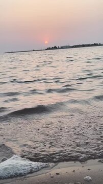 Vertical mobile video of beach sunset with gentle waves washing over sand and sea foam, Vertical POV shot of a relaxing beach evening and distant hazy shoreline