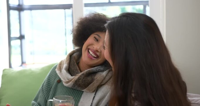 Diverse female friends with scarf laughing, leaning on couch by window, sharing smartphone and mug