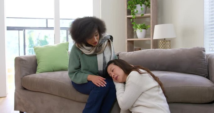 Diverse female friends: Asian woman seeking comfort, seated woman softly patting head on gray sofa