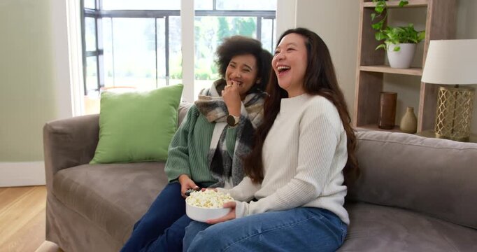 Two women watching show starting on gray couch by window, laughing, nibbling popcorn, left pointing
