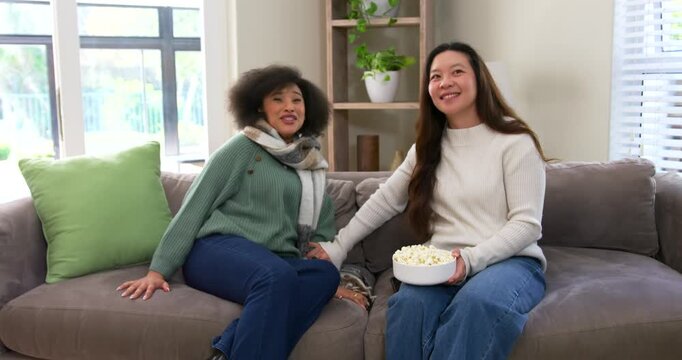 Diverse female friends reacting to off-frame sound, holding popcorn bowl and grasping hands on sofa