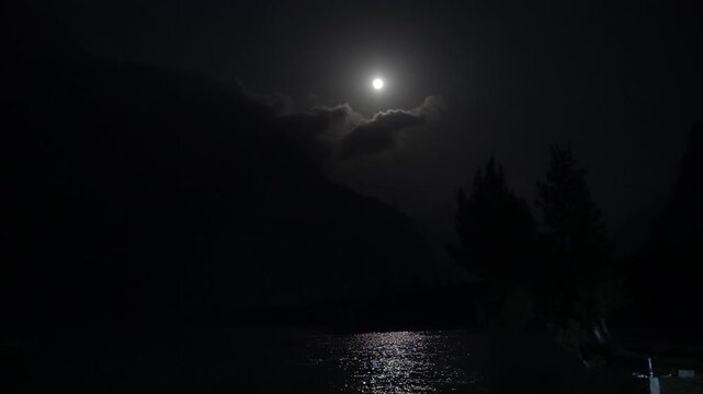 Full moon shines above a dark mountain lake, casting a bright reflection across the water