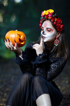 Halloween beauty portrait of woman in forest with Halloween pumpkin in hand. Santa Muerte makeup.