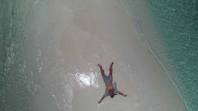 Aerial pullback from relaxed man lying on the famous pristine sandbar of Cayo de Agua Island, surrounded by turquoise Caribbean waters of Los Roques National Park, Venezuela.