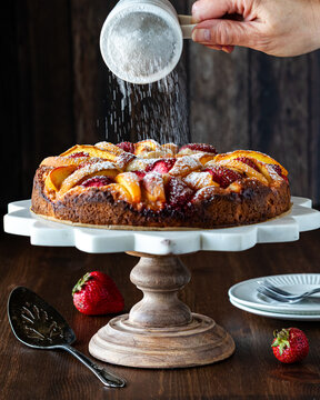 A hand shaking powdered sugar onto a homemade peach cake causing motion blur.