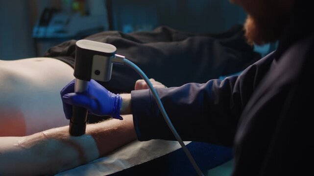 Close up of professional male therapist using massage gun on athlete body hand and leg muscles. Modern physiotherapy and sports recovery session in clinic, deep tissue percussion therapy concept.