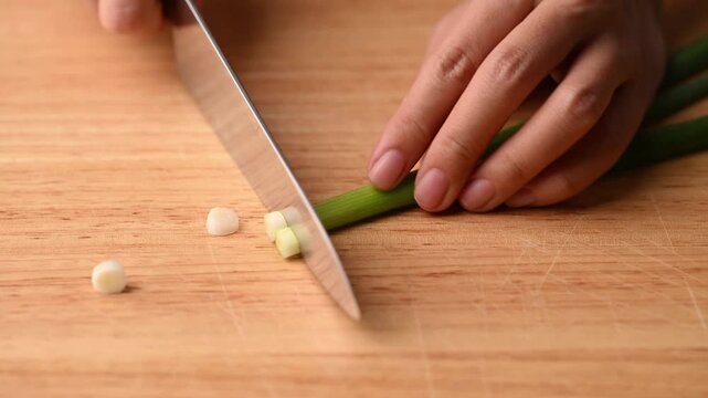 Close-up of woman's hands chopping spring onion with a knife on a cutting wooden board, preparing ingredients for cooking