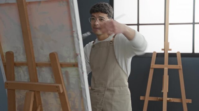 Young painter in apron wearing glasses, brush in hand, arm reaching to paint on canvas at easel by window in studio; quiet concentration.