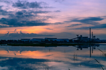 Fototapeta premium Landscape of factory industry buildings with dark blue and orange sunset sky reflection on water in the river. Warehouse building at night. Clean environment around factory. Factory closed.