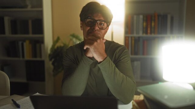Man wearing glasses resting chin on clasped hands over laptop and books in study building at night; late night focus.