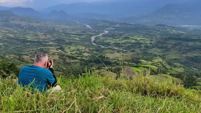 Rear view of male photographer sitting on grassy hill taking photos of vast green mountain valley landscape with winding river during outdoor travel wellness adventure
