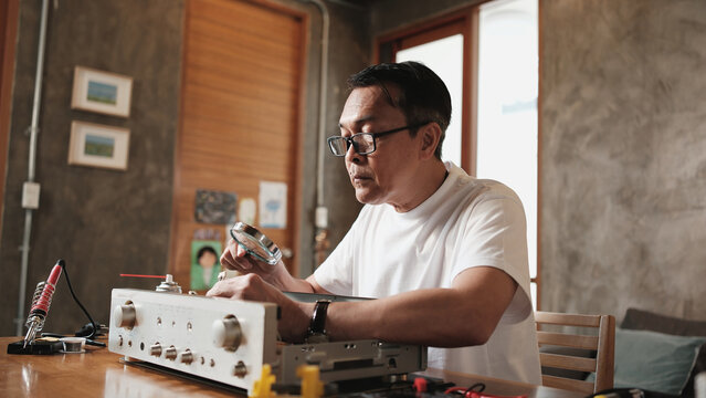Mature Asian man disassembling and checking audio equipment, looking with magnifying glass at circuit board details for repair, home appliance DIY fix, electronics component hobby, handyman workshop.