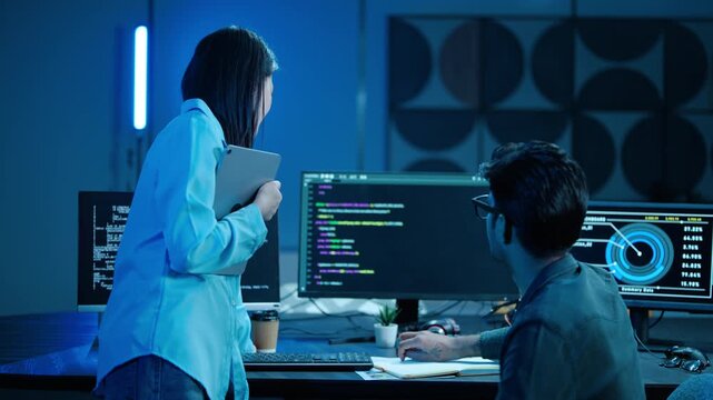 Tech professionals analyzing software code on multiple desktop monitors in a dark office. Male programmer and female manager collaborating late at night on complex cybersecurity data dashboards.